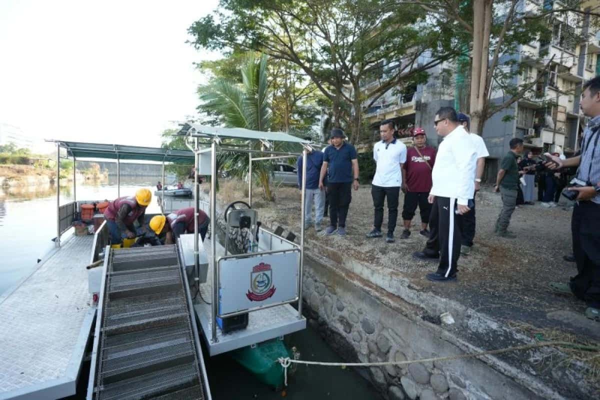 Petugas berhelm kuning berada di kapal pembersih limbah, sementara Munafri dan rombongan berdiri di tepi perairan
