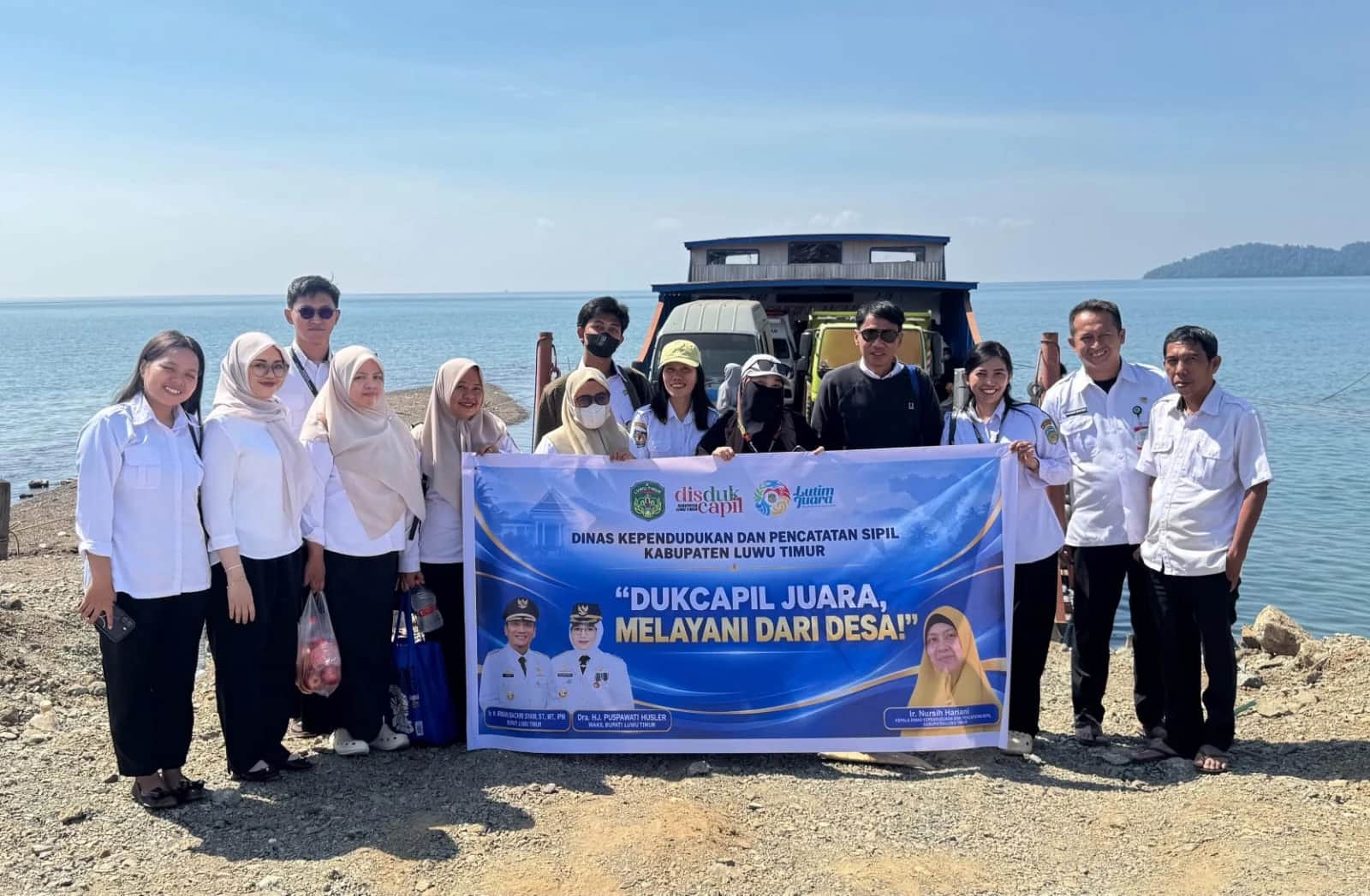 Tim Dukcapil Luwu Timur memegang spanduk layanan desa di tepi danau dengan perahu di belakang mereka